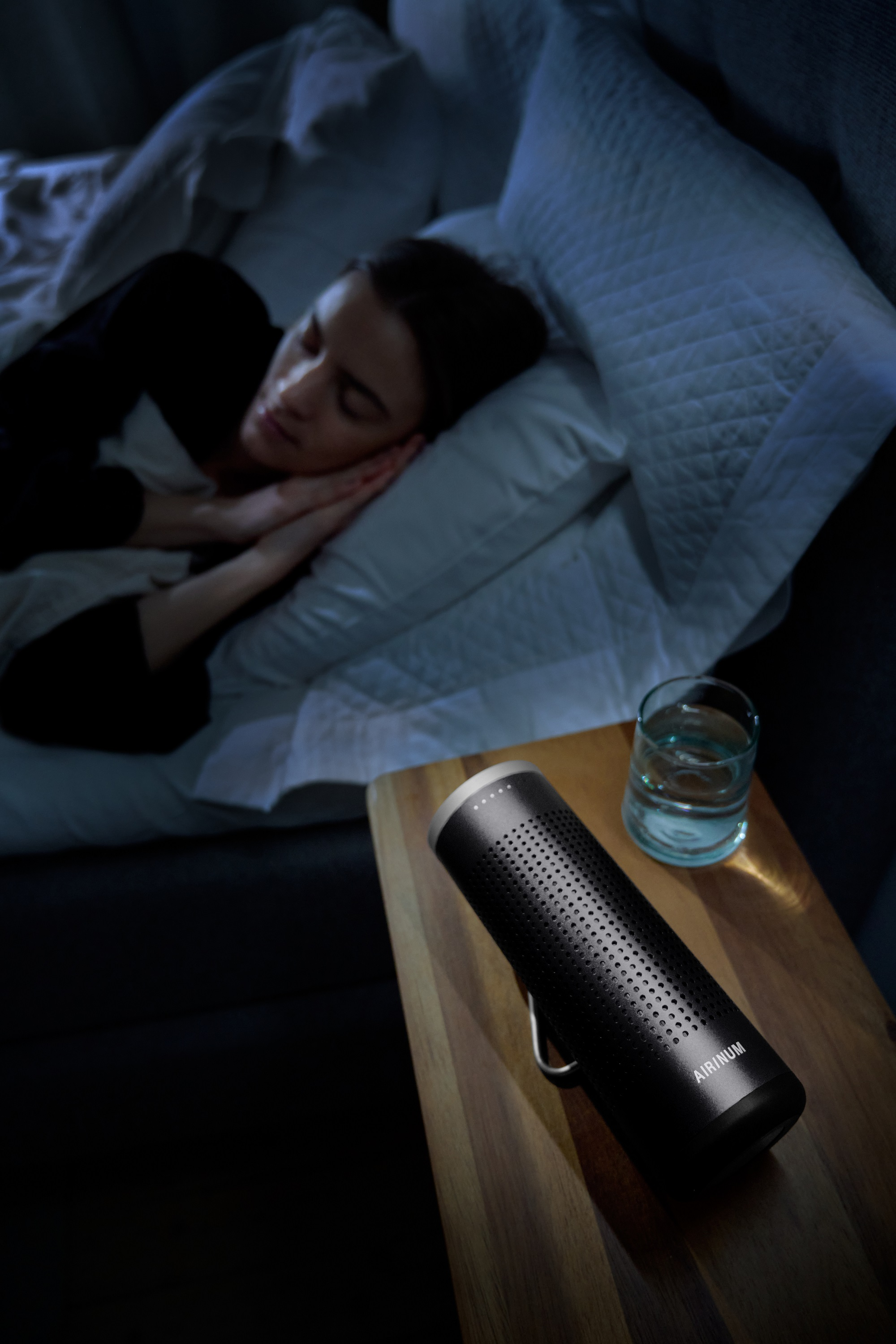 Eine Frau schläft im Bett neben einem Luftreiniger auf einem Holztisch, ein Glas Wasser steht ebenfalls auf dem Tisch.