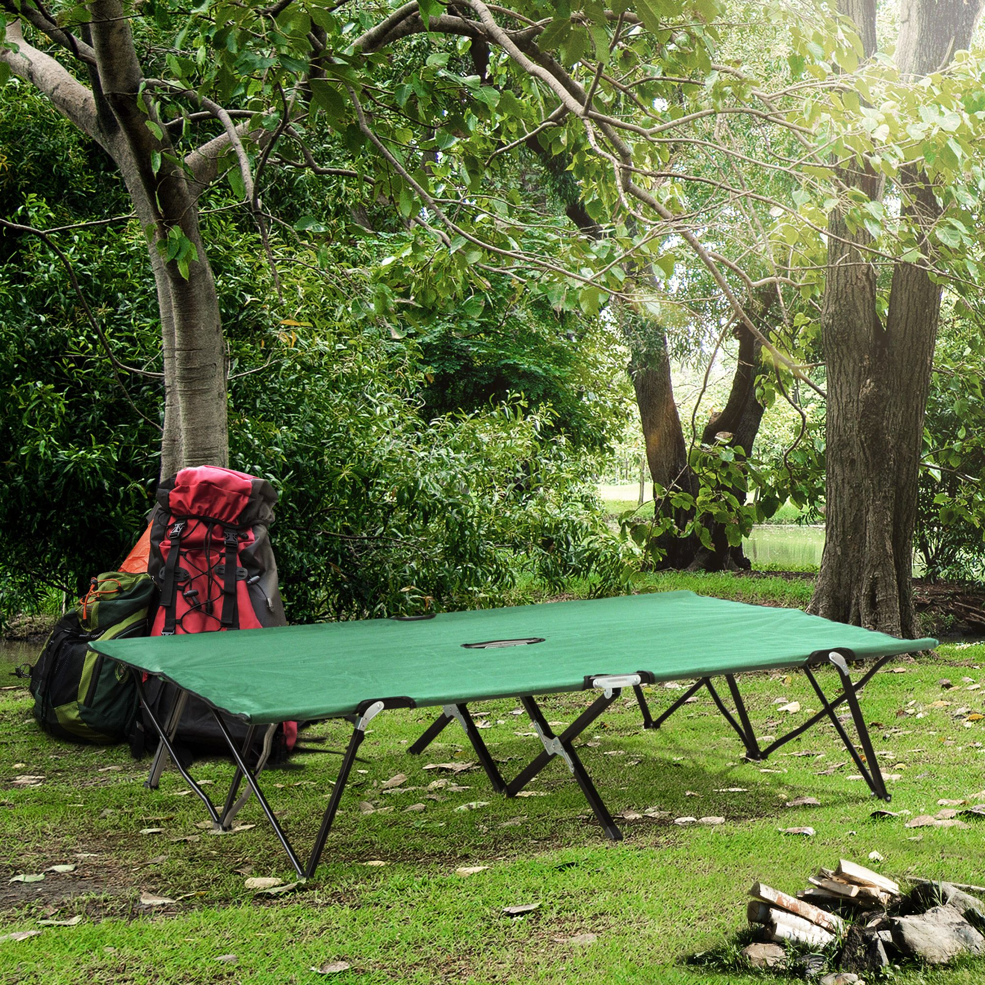 Grüne Campingliege auf einer Wiese, mit Bäumen, Rucksäcken und einem kleinen Holzstapel im Hintergrund.