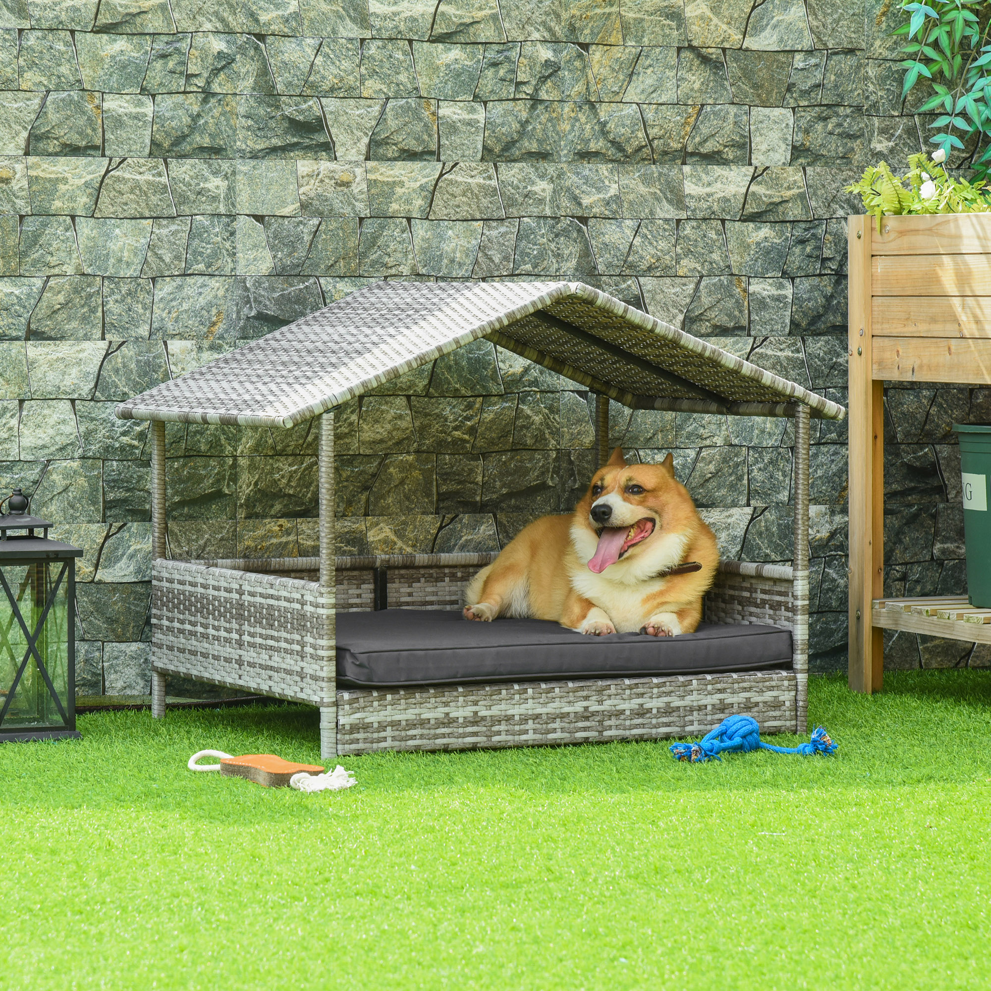 Hundebett draußen. Ein Hund liegt auf dem Bett unter einem Dach. Gras, eine Steinmauer und ein Spielzeug sind sichtbar.