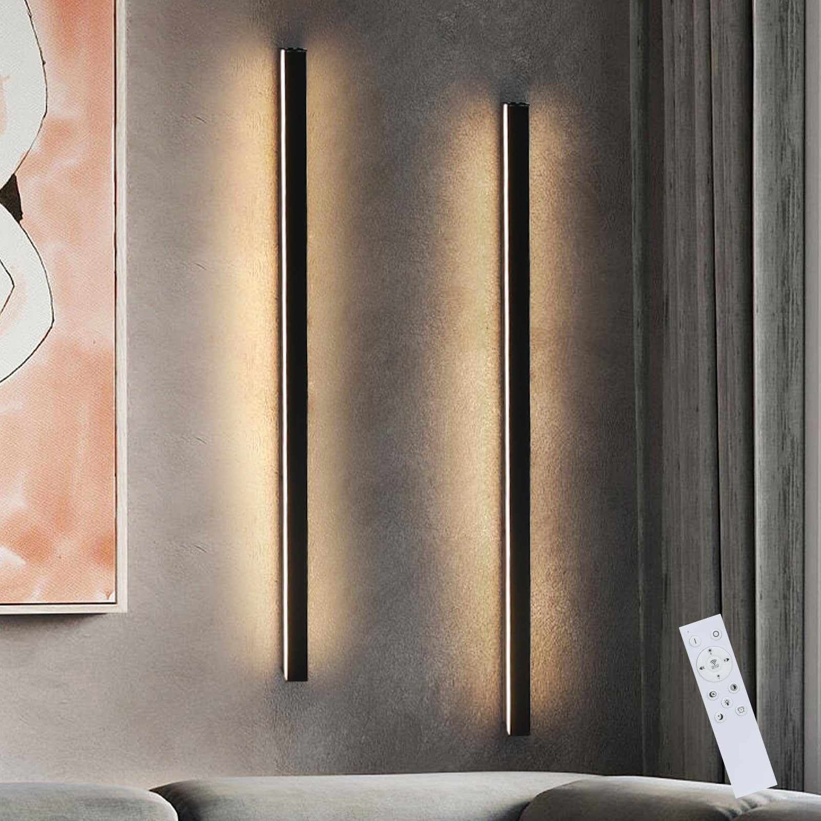 Due luci a LED nere verticali su un muro grigio, con un dipinto a sinistra e un telecomando a destra.
