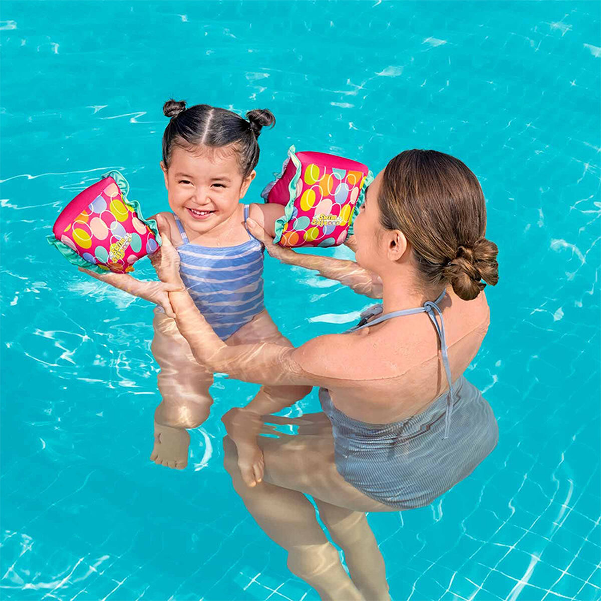 Una donna e una bambina in piscina con braccioli. La bambina sorride. I braccioli sono rosa e gialli.