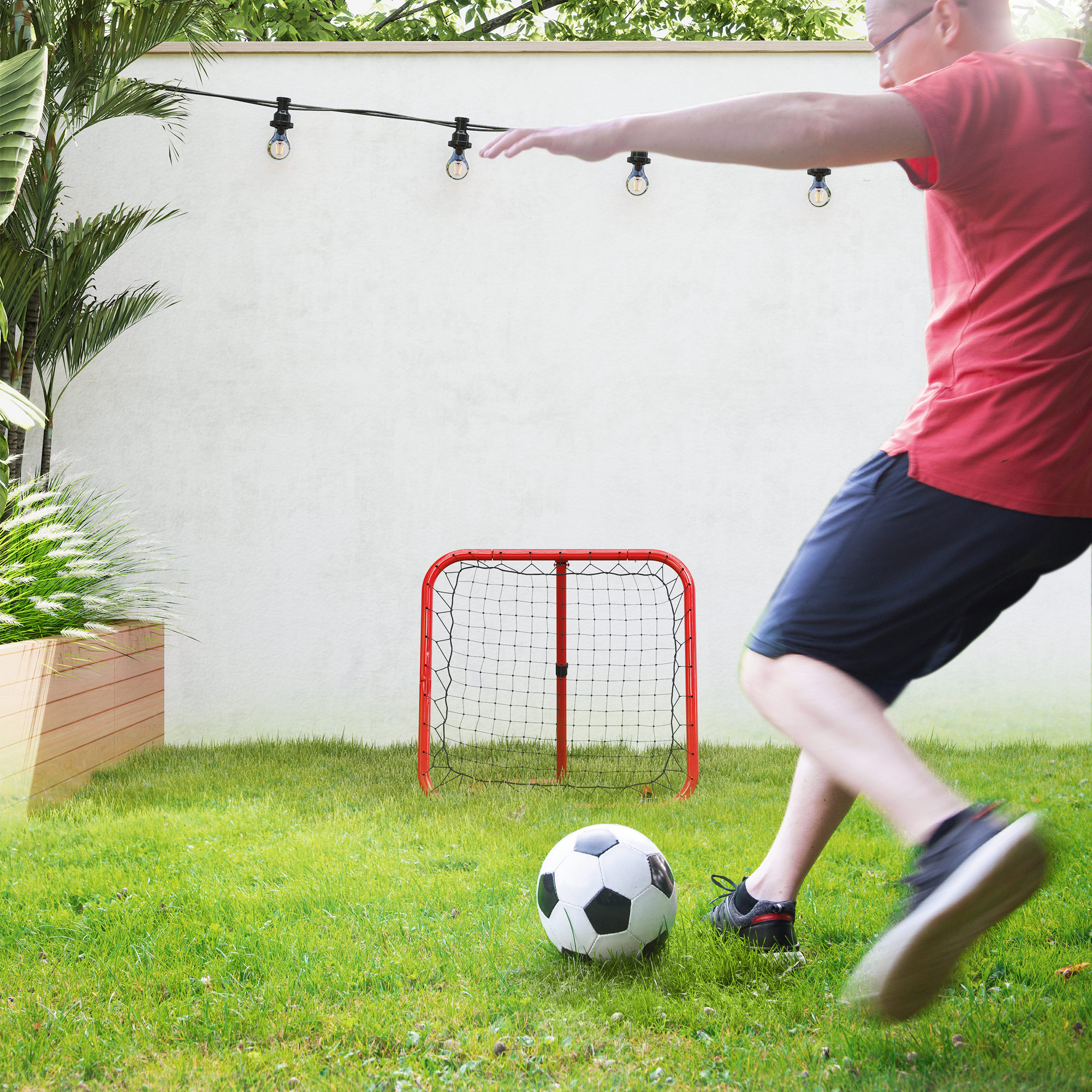 Ein Mann tritt einen Fußball in Richtung eines roten Netzes in einem Hinterhof mit Lichterketten.