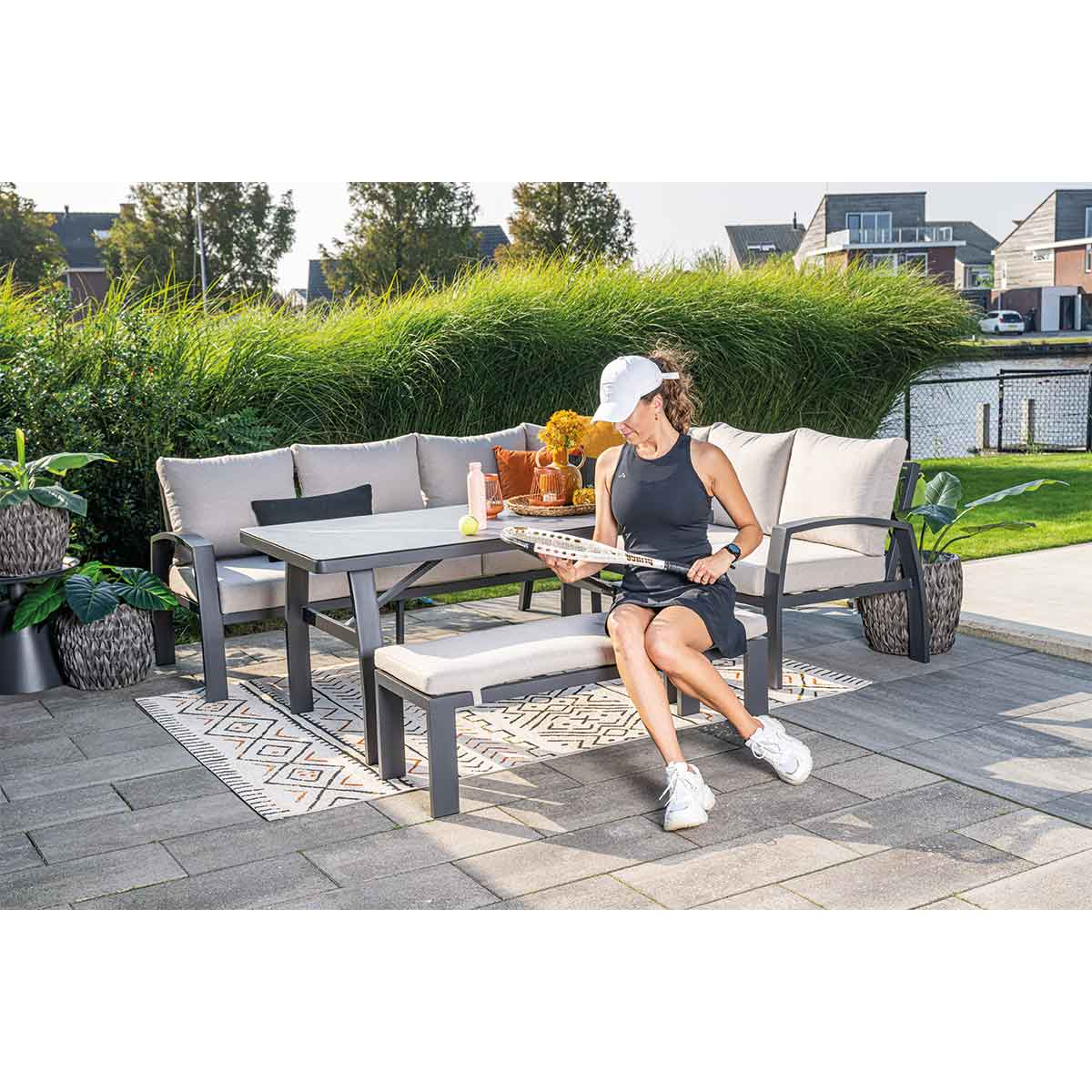 Vrouw zit op tuinmeubilair, houdt tennisracket vast. Tafel, vloerkleed en planten zijn zichtbaar.