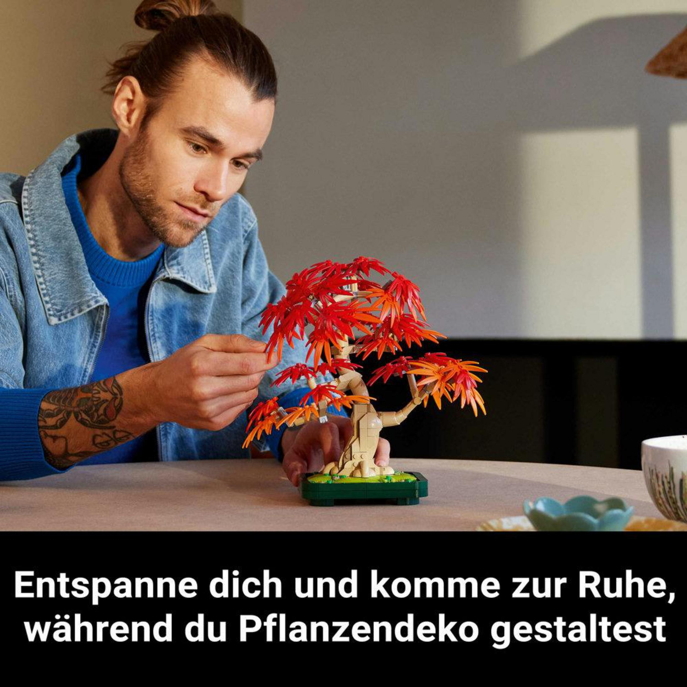 Kırmızı ve turuncu yapraklı bir Lego bonsai ağacı yapan bir adam, iç mekanda.