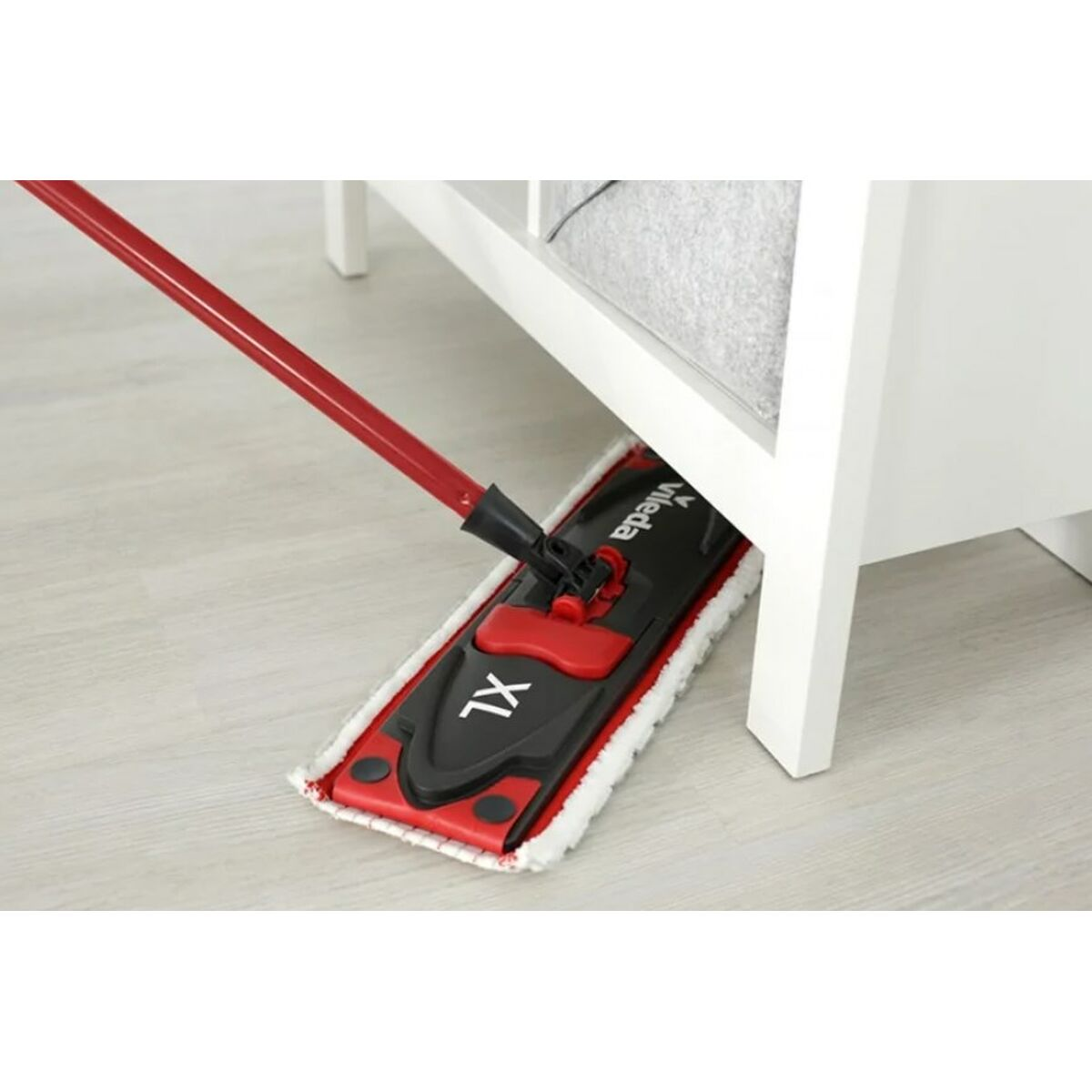 Mocio Vileda XL rosso e nero che pulisce sotto un mobile bianco, su pavimento in legno chiaro.