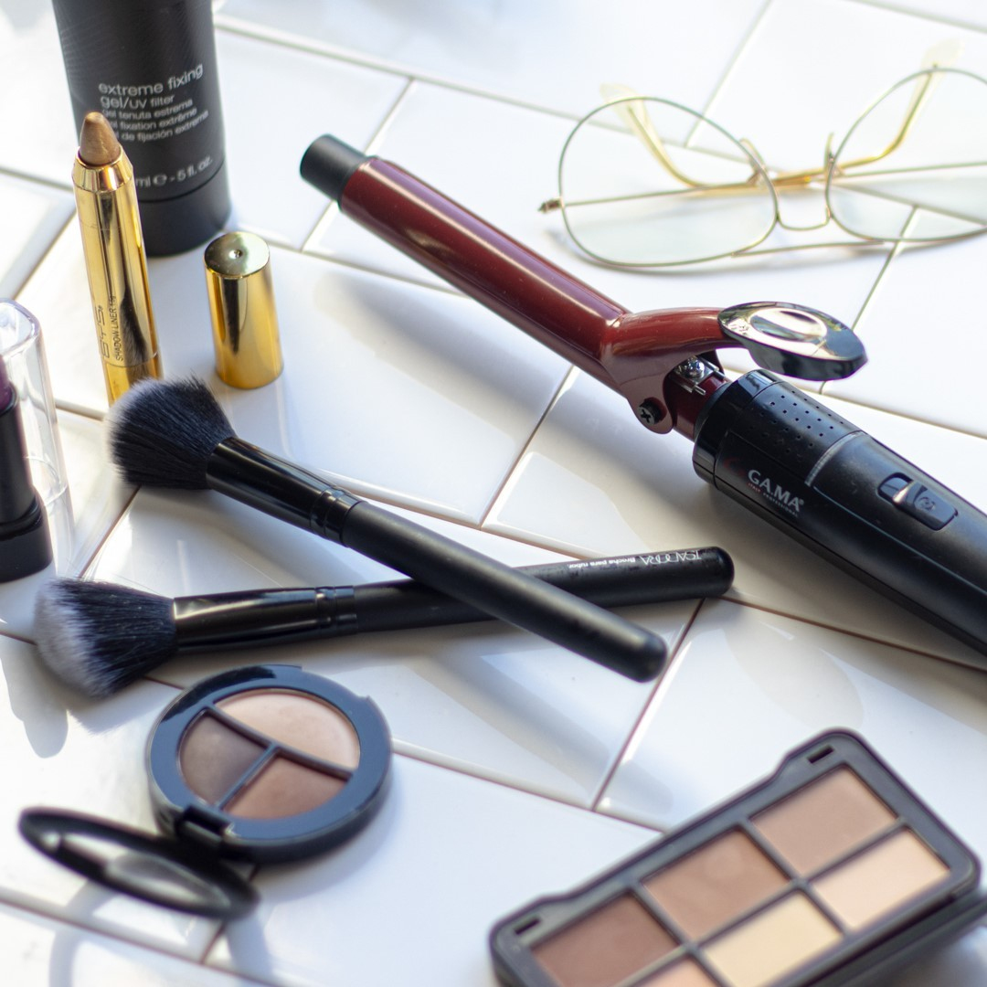 Prodotti per il trucco su una superficie di piastrelle bianche. Include pennelli, ombretto e arricciacapelli.