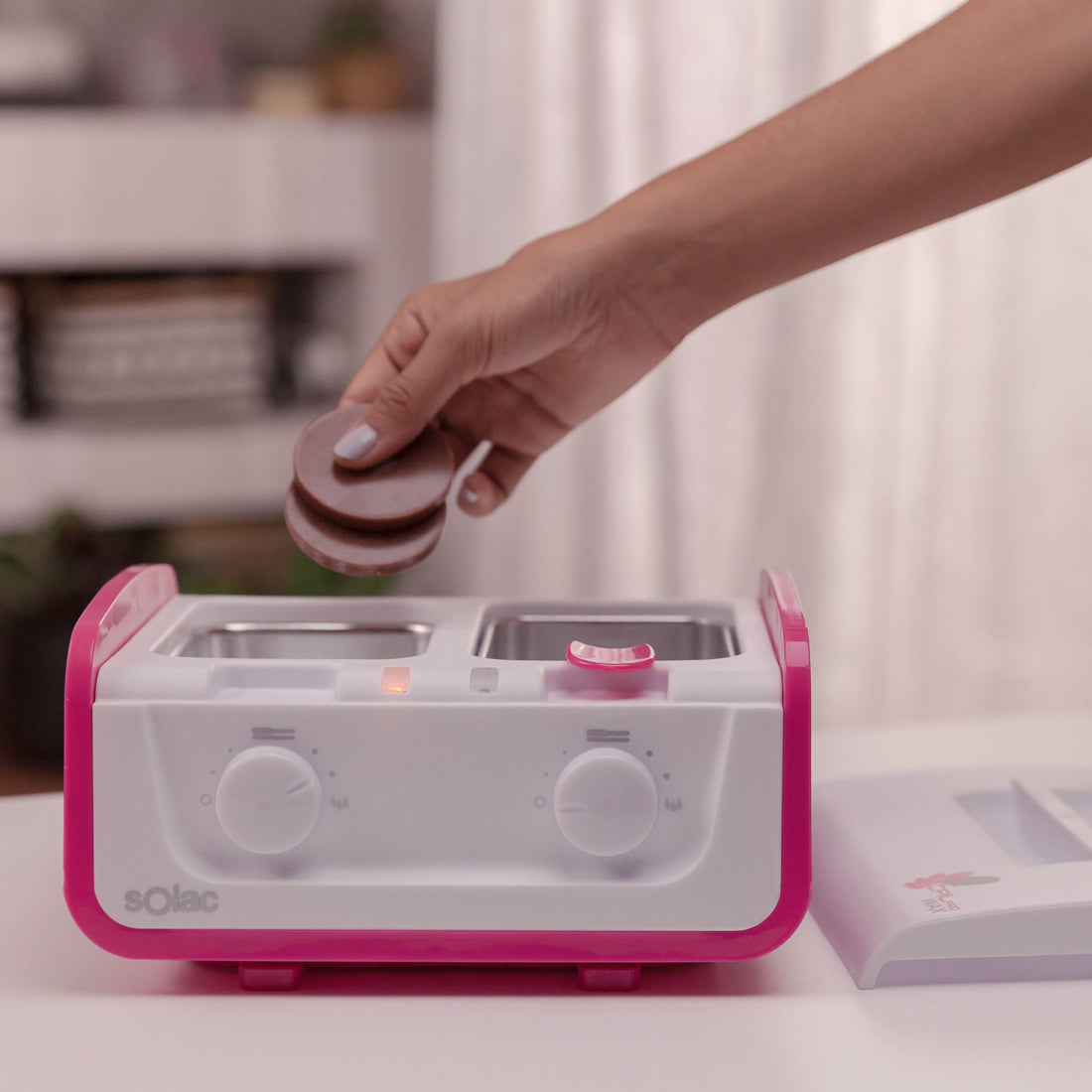 Hand legt Wachsplatten in einen rosa-weißen Wachserhitzer, mit weißem Hintergrund.