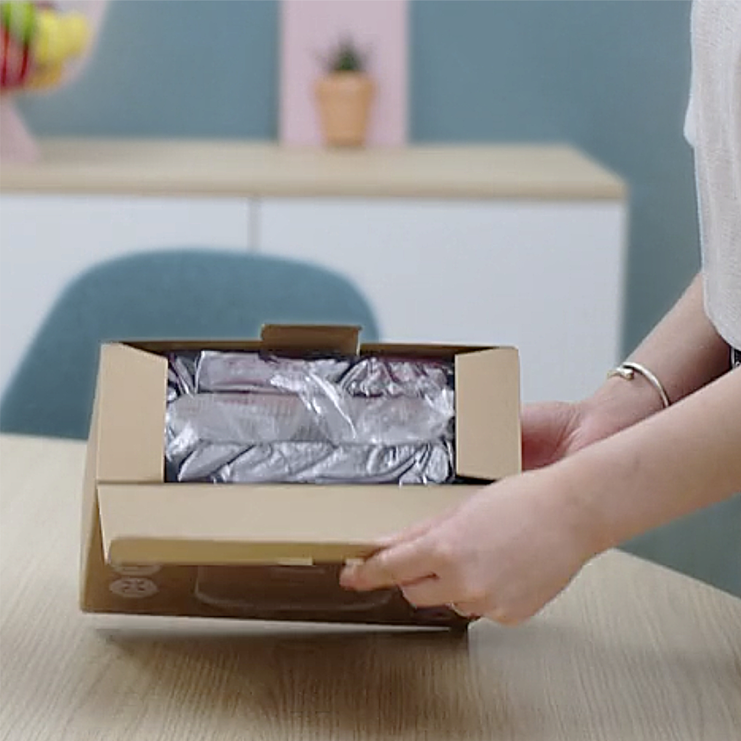 Person öffnet Pappschachtel mit verpackten Gegenständen auf einem Holztisch.