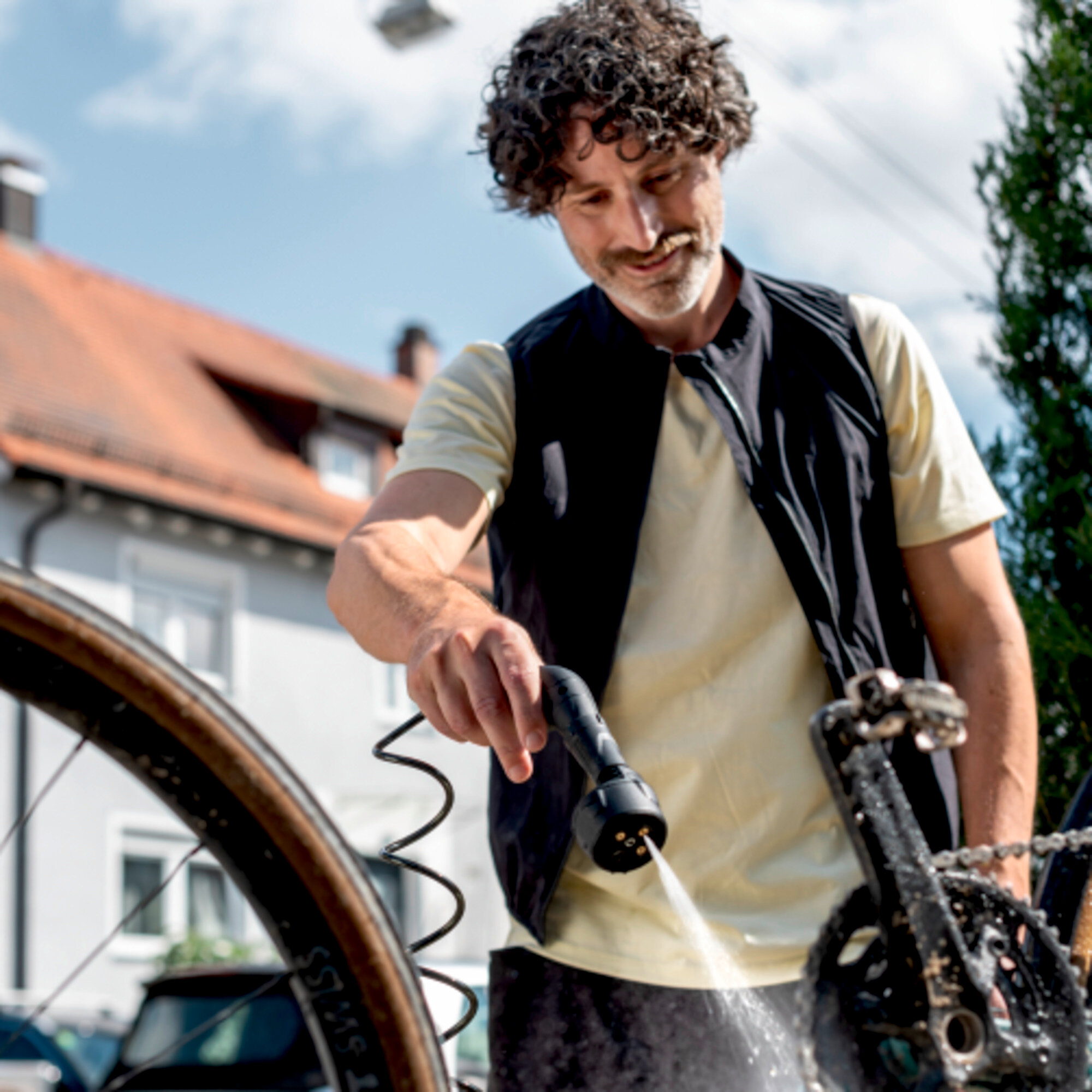 Een man wast een fiets met een hogedrukreiniger, een wit huis op de achtergrond.
