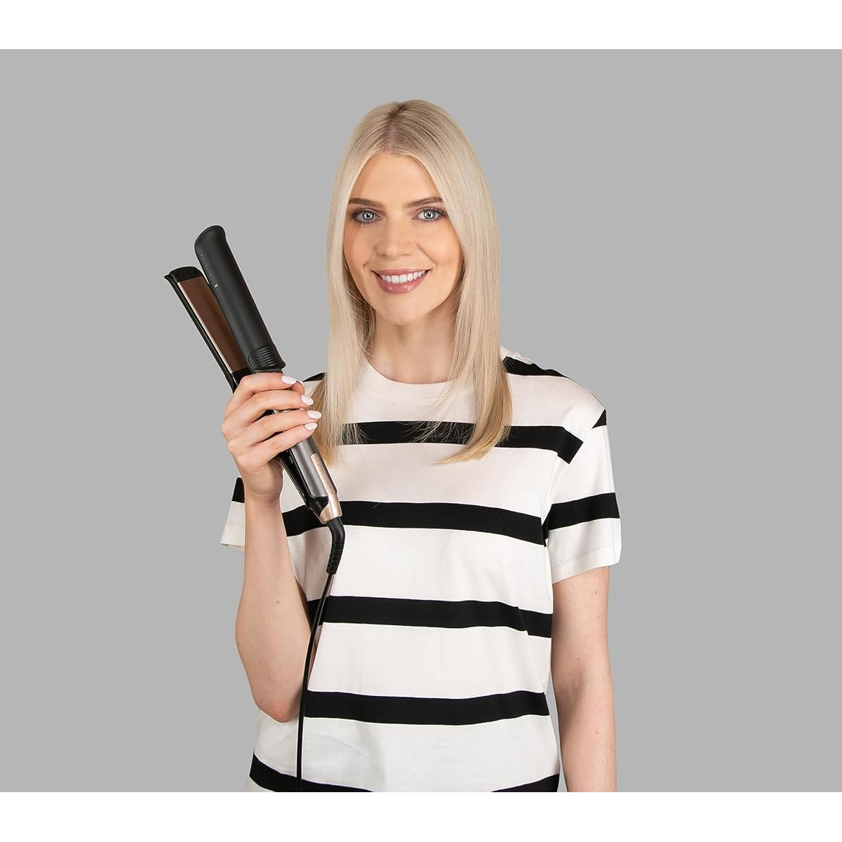 Eine Frau mit blonden Haaren hält einen Haarglätter und lächelt in die Kamera.