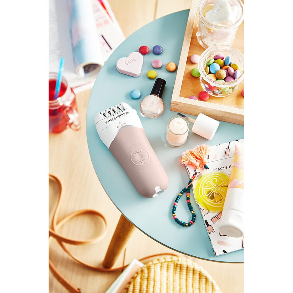 Een epilator en diverse schoonheidsartikelen liggen op een blauwe tafel. Een drankje op de achtergrond.