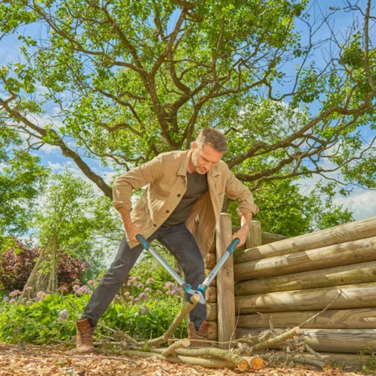 Mężczyzna używający nożyc Gardena do cięcia gałęzi. Zielone drzewa, drewniane ogrodzenie i niebieskie niebo w tle.