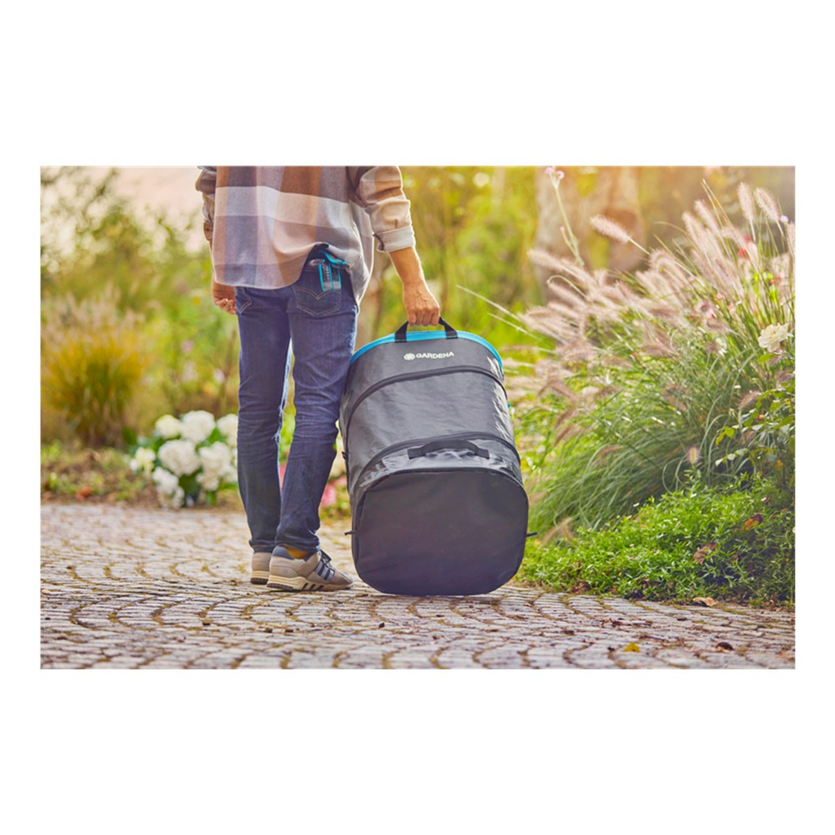 Osoba niesie złożoną czarno-niebieską torbę Gardena na kamiennej ścieżce w ogrodzie.