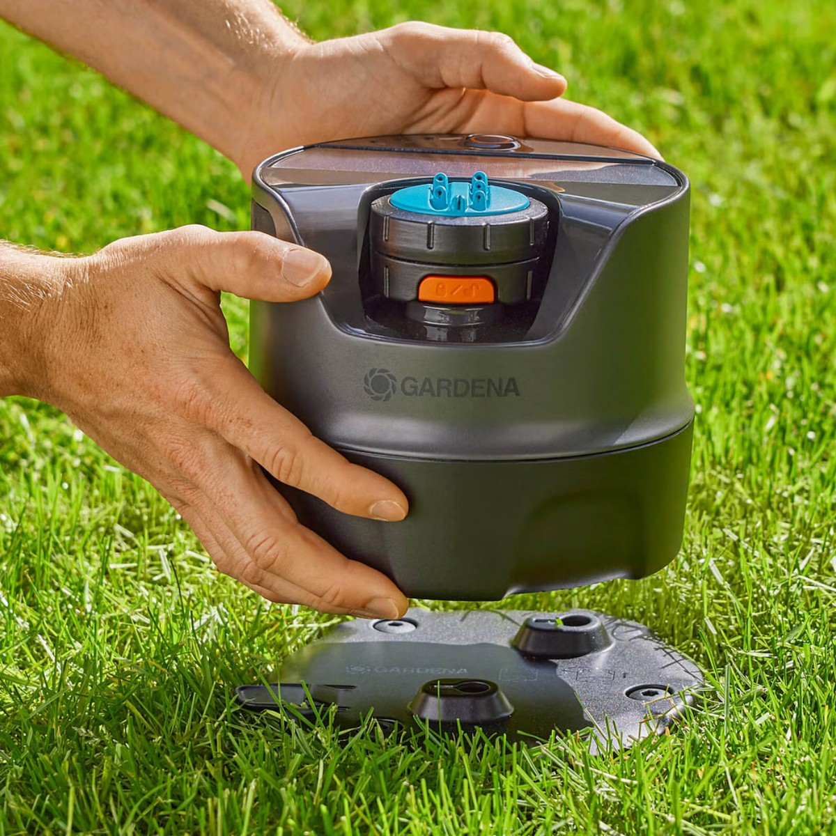 Le mani posizionano una base per irrigatore nera a terra. Il dispositivo ha una parte superiore blu, arancione e nera.