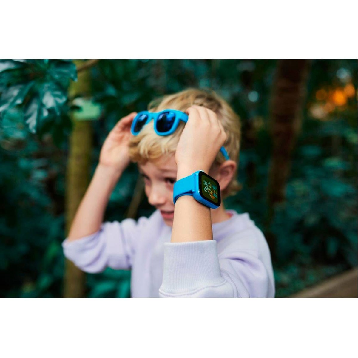 Een jongen met een blauwe smartwatch en zonnebril, omringd door groen.