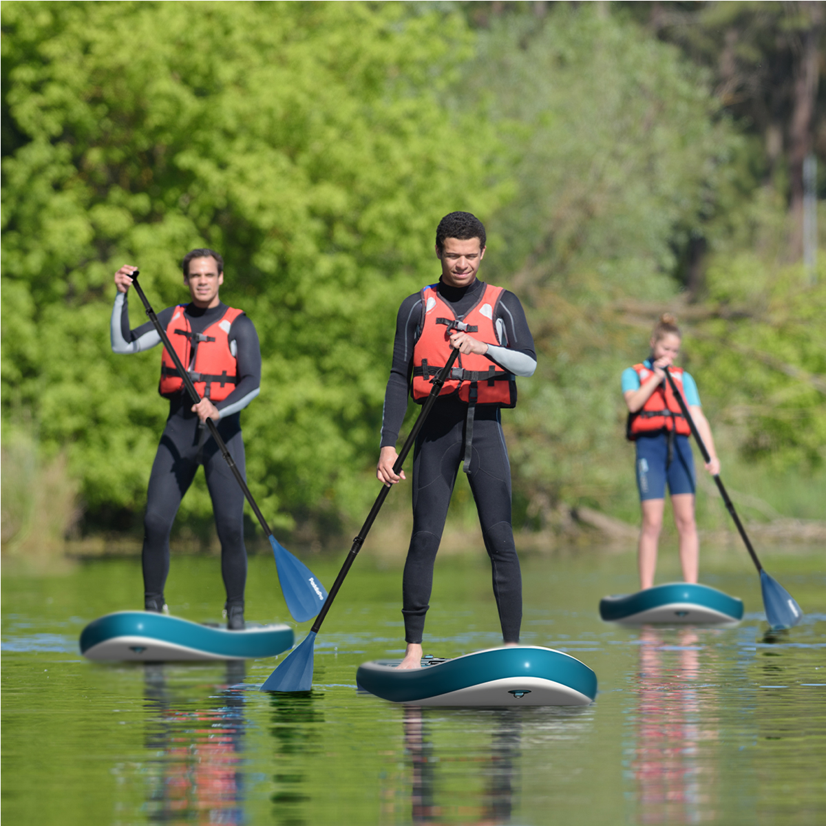 Trois personnes font du paddleboard sur l'eau. Ils portent des combinaisons et des gilets de sauvetage. Arbres verts en arrière-plan.