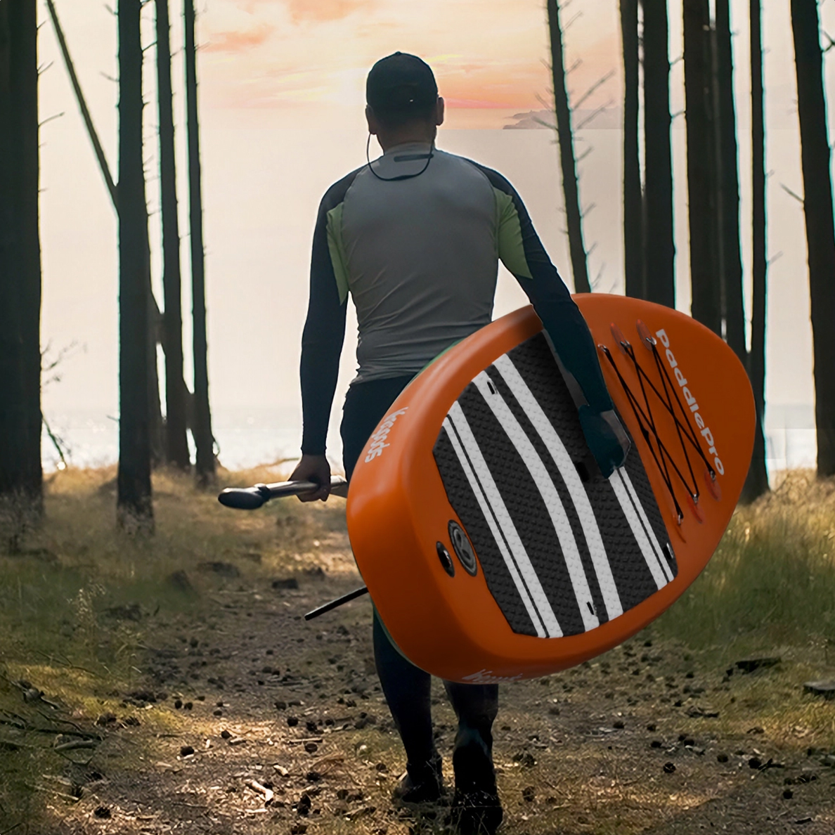 Ein Mann trägt ein orangefarbenes SUP-Board und Paddel durch einen Wald bei Sonnenuntergang.