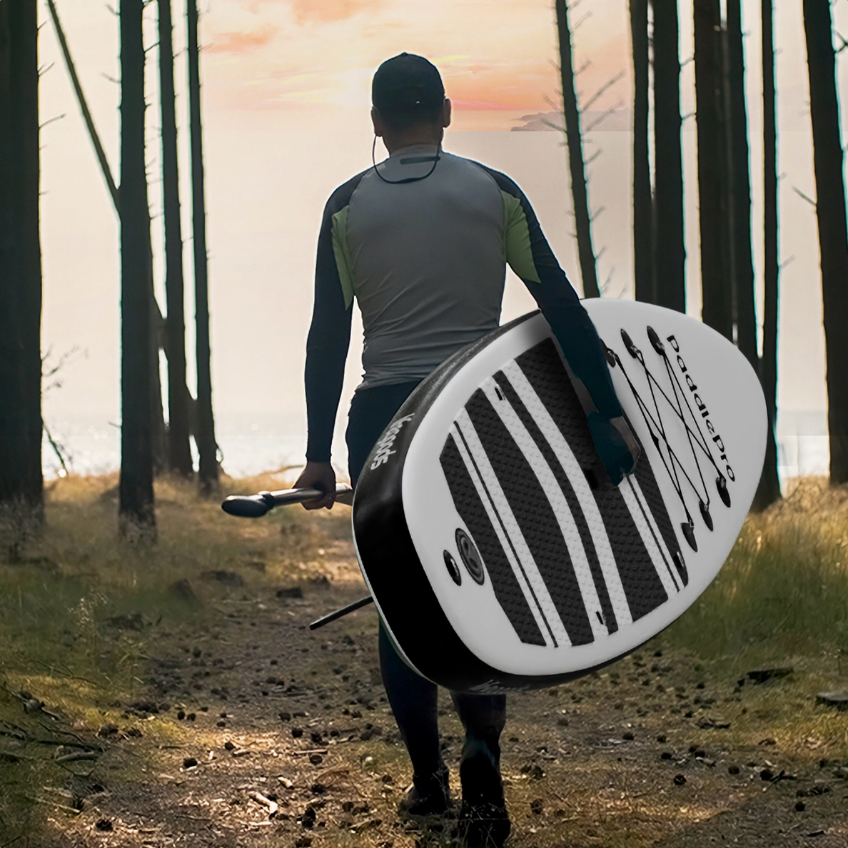 A man walks on a path through a forest, carrying a paddleboard and paddle over his shoulder.