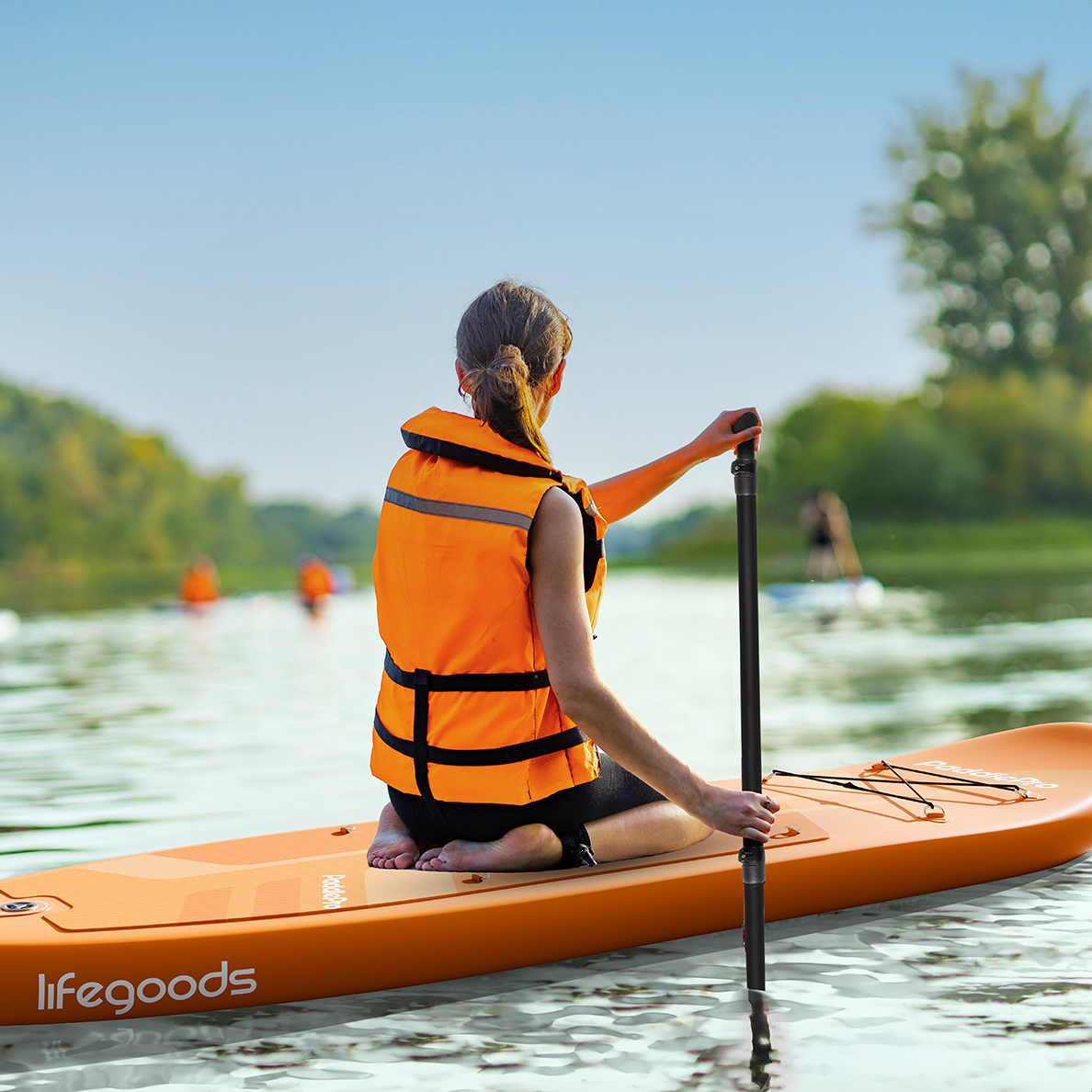 Vrouw in een oranje zwemvest aan het suppen op een rivier. Ze knielt en houdt een peddel vast.