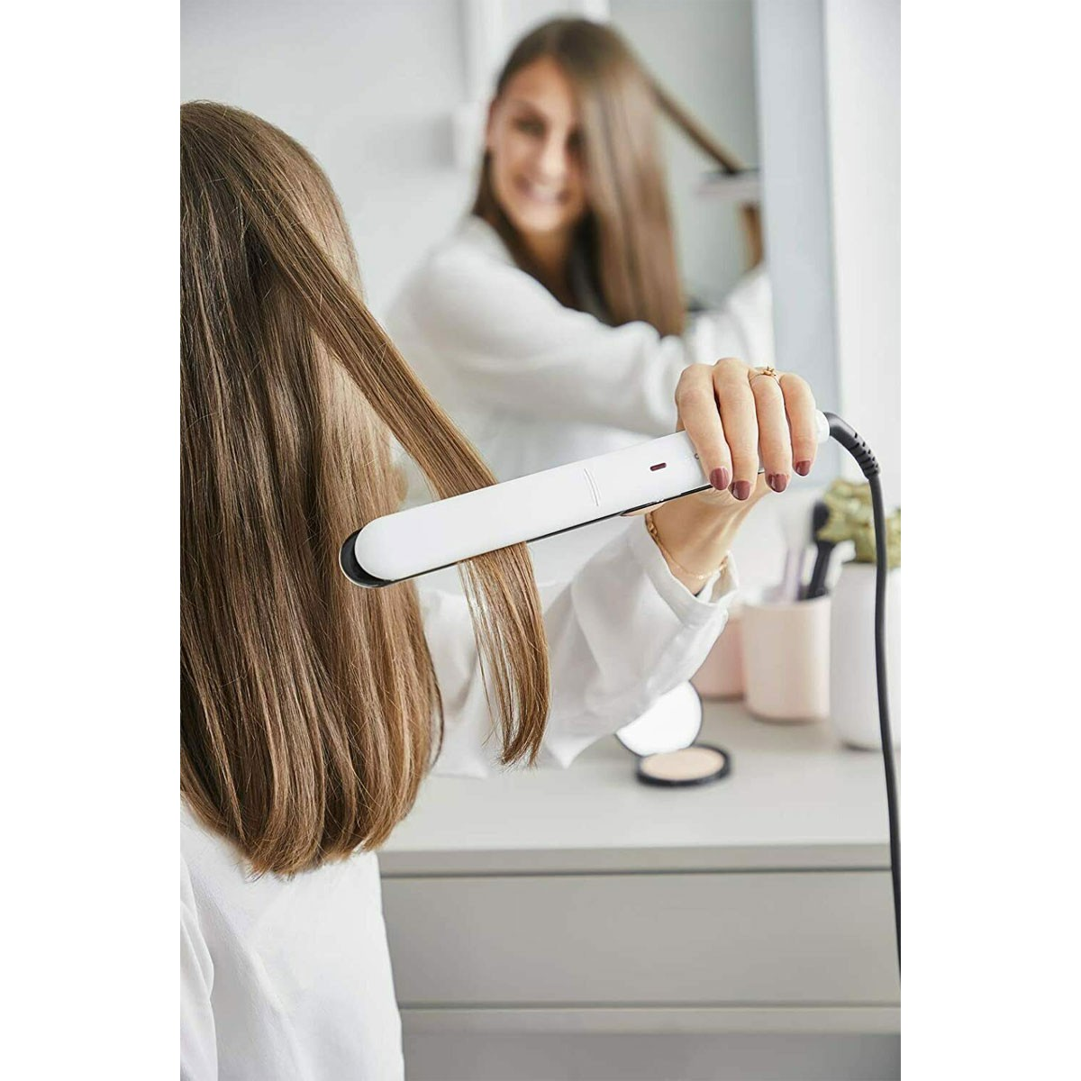 Donna che usa la piastra per capelli davanti a uno specchio. Sta sorridendo, concentrata sui suoi capelli.