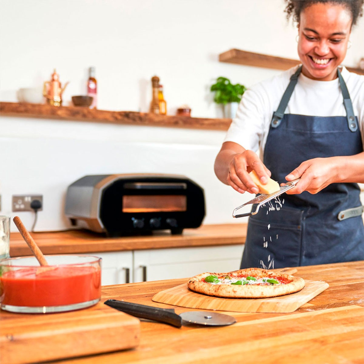 Eine Person, die Käse auf einer Pizza reibt. Ein Holzofen ist im Hintergrund. Pizza auf dem Holzbrett.