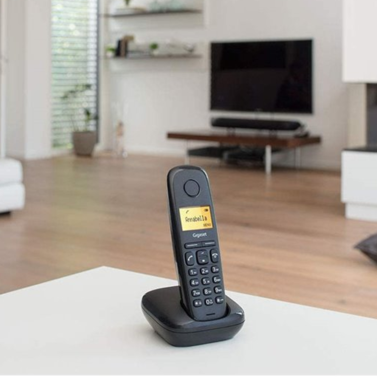 Un teléfono negro en una base negra sobre una mesa blanca en una sala de estar. Un televisor en el fondo.