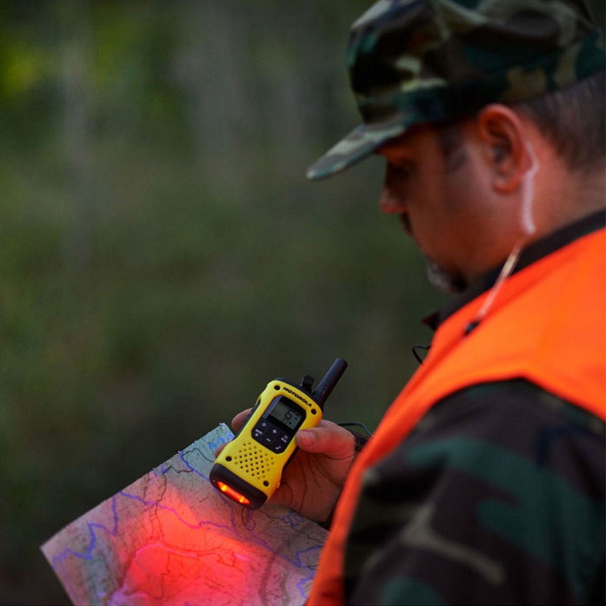 Mężczyzna w kamuflażu trzyma żółte krótkofalówki i mapę z podświetlonym obszarem.