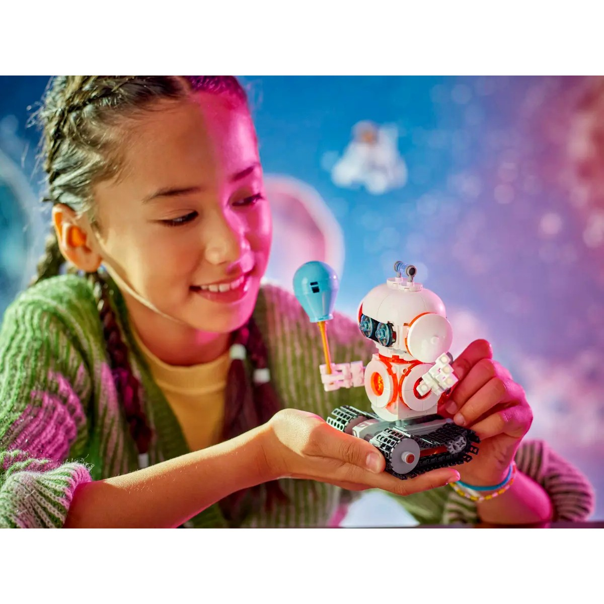 Dziewczynka uśmiecha się, trzymając robota Lego z niebieskim balonem. Widoczne jest kolorowe tło.