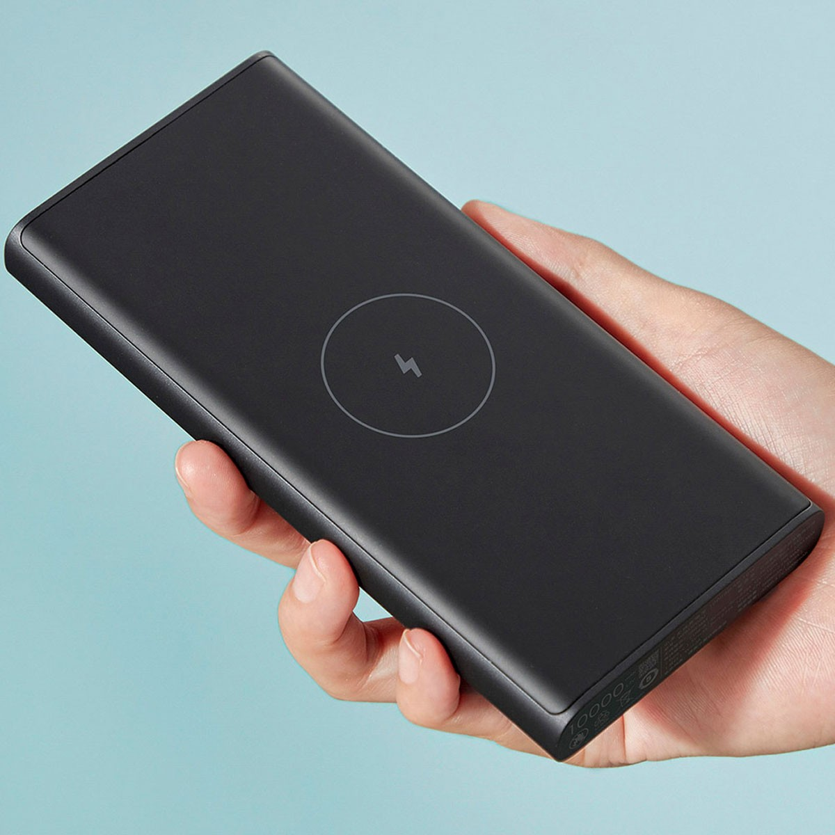Power bank nero tenuto in mano, con simbolo di ricarica wireless. Sfondo azzurro chiaro.