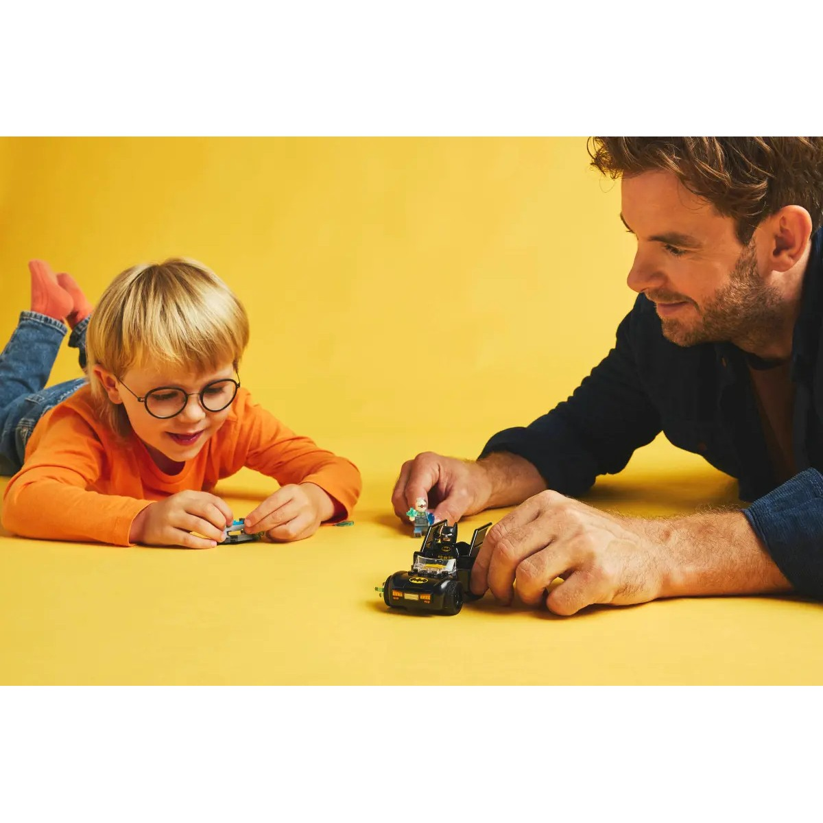 Un ragazzo e un uomo che giocano con un'auto Lego Batman su sfondo giallo.