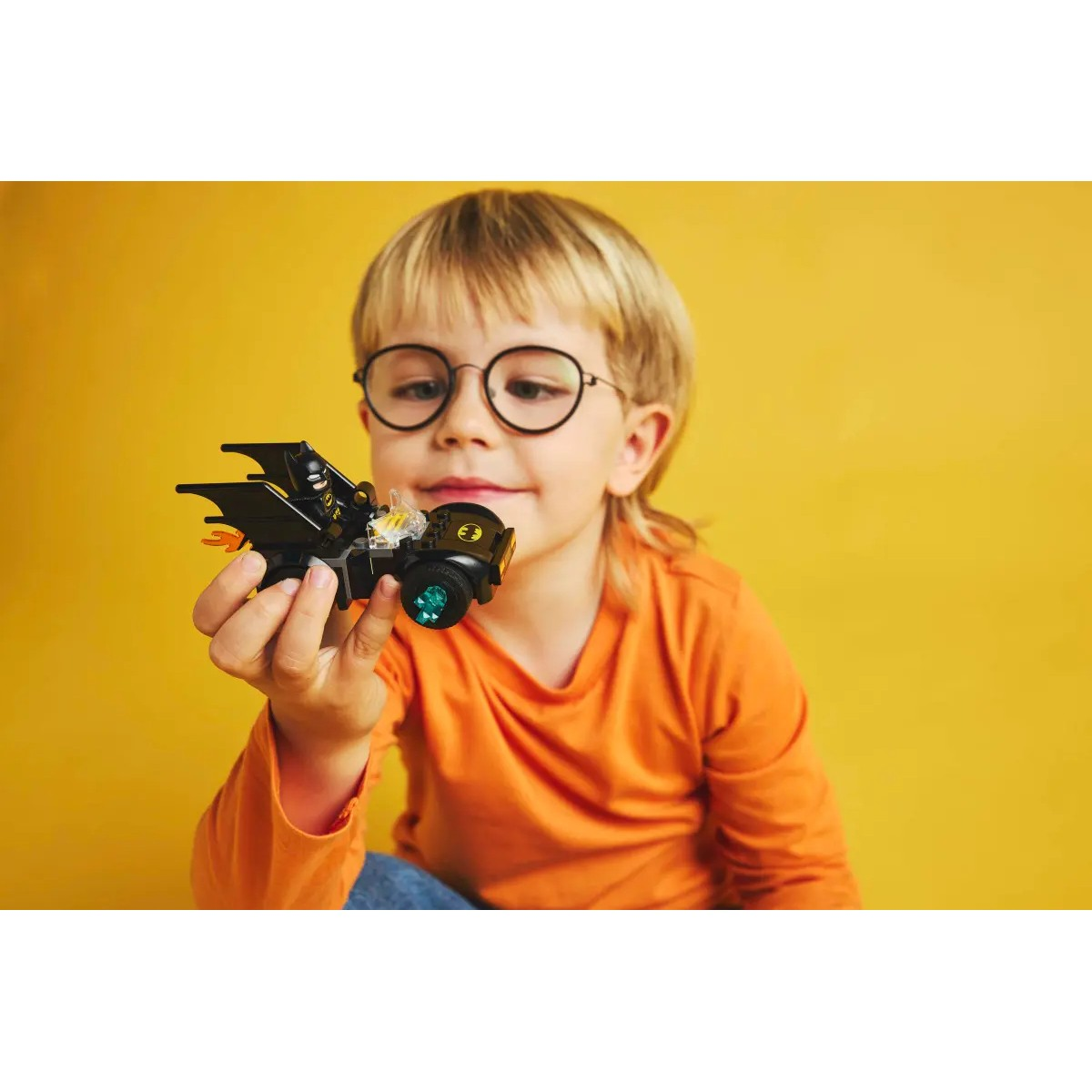 Un bambino in arancione tiene in mano un'auto Lego Batman, guardandola con gli occhiali.