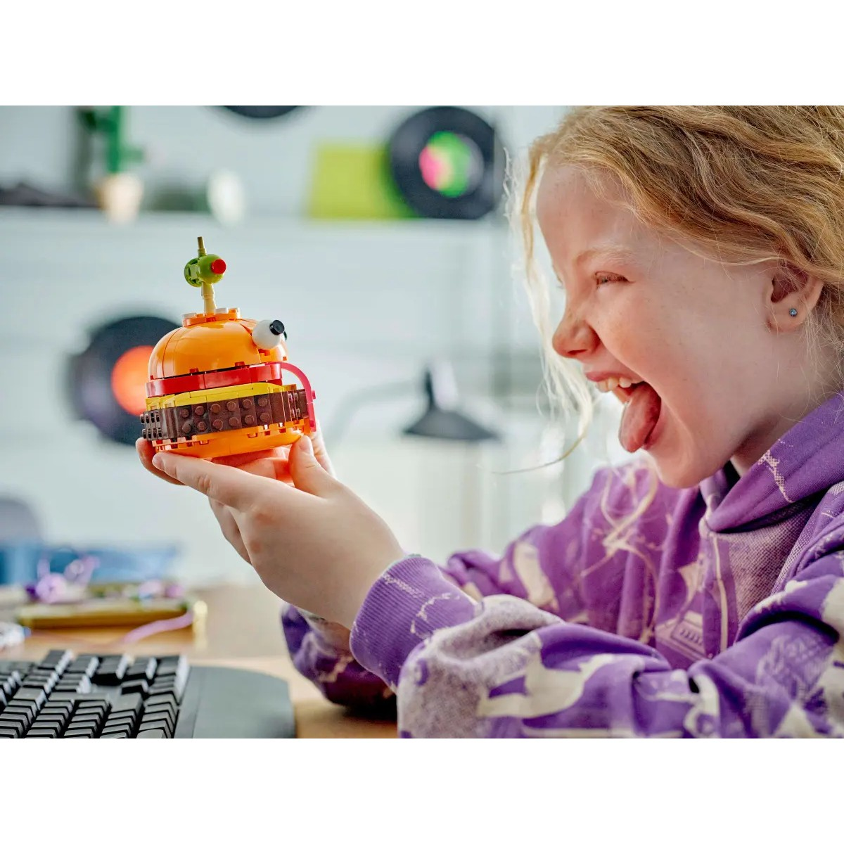 Kind hält Lego Burger, Zunge raus, Tastatur im Hintergrund.