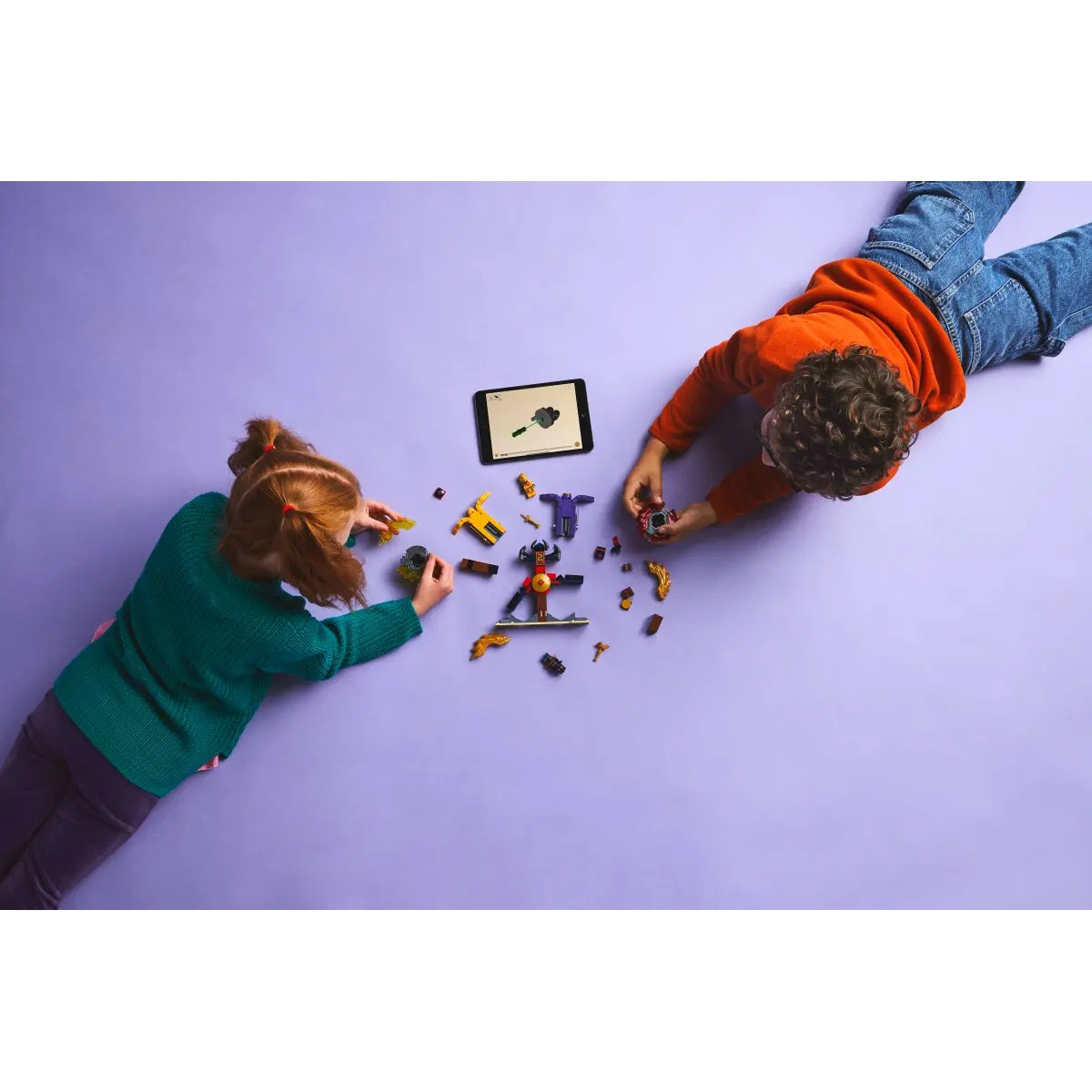 Dos niños tumbados en una superficie morada jugando con juguetes Lego. Una tableta está en el medio.