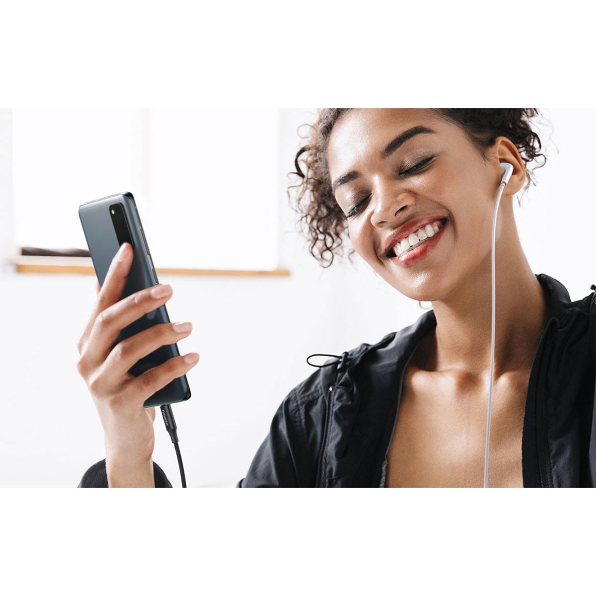 Uśmiechnięta kobieta trzyma telefon ze słuchawkami, słuchając muzyki.