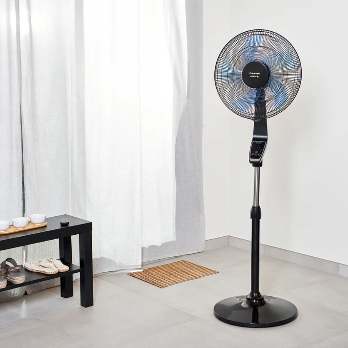 Czarny wentylator stojący w pokoju. Za nim biała zasłona i czarny stojak na buty.