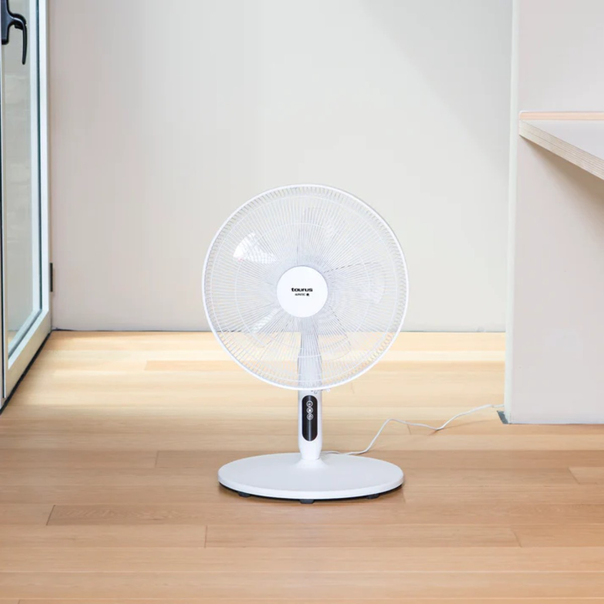 Biały wentylator podłogowy stoi na drewnianej podłodze. Tło to biała ściana i drzwi.