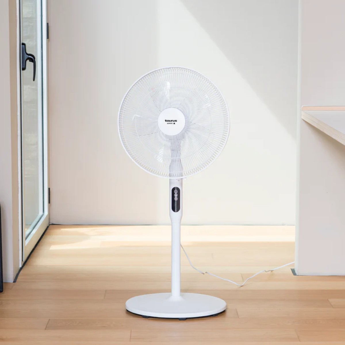 Biały wentylator podłogowy stoi na drewnianej podłodze. Tło to biała ściana i drzwi.