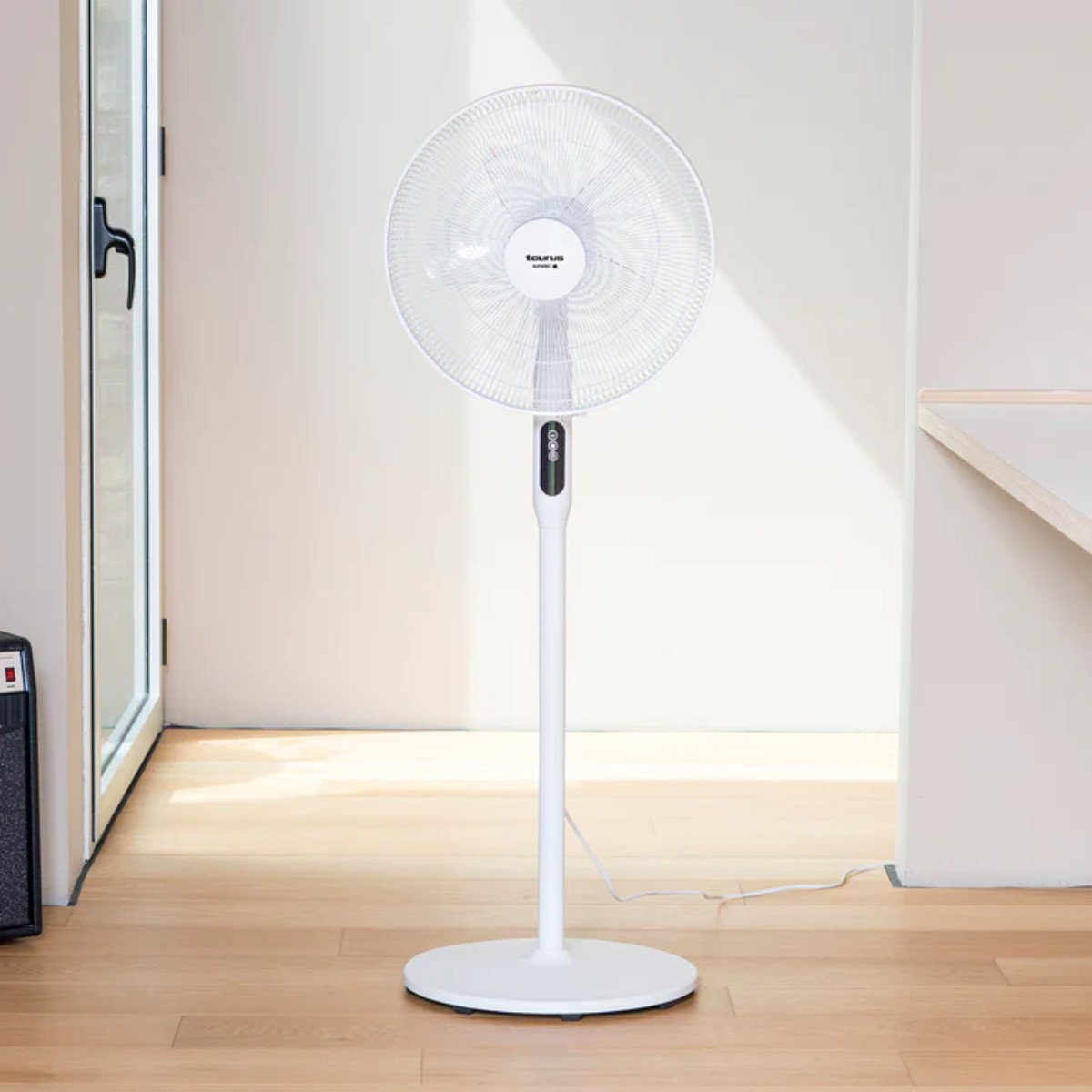 Biały wentylator podłogowy stoi na drewnianej podłodze. Tło to biała ściana i drzwi.