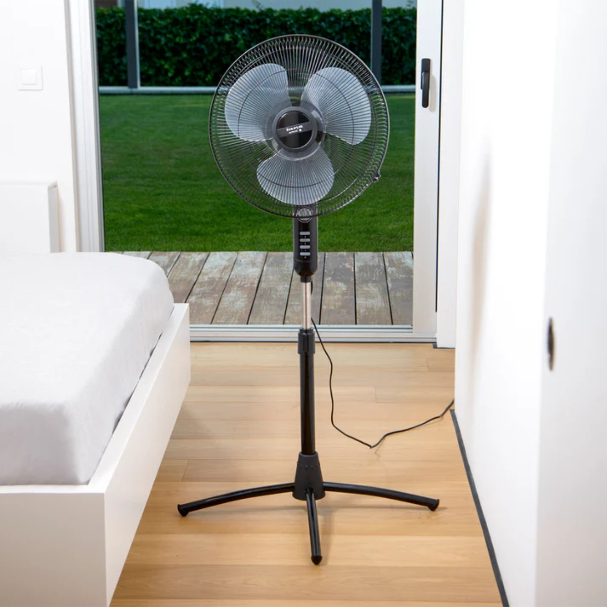 Czarny wentylator stojący w pokoju z łóżkiem i oknem. Widoczne są łopatki wentylatora.