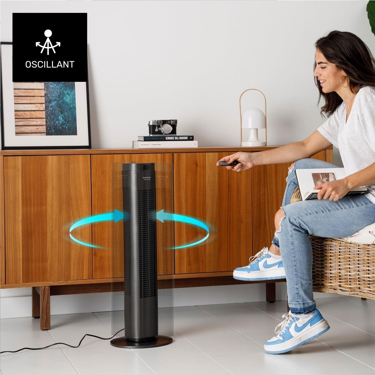 Kobieta siedzi, wskazując na pilota, wysoki, czarny wentylator pracuje obok drewnianej szafki.