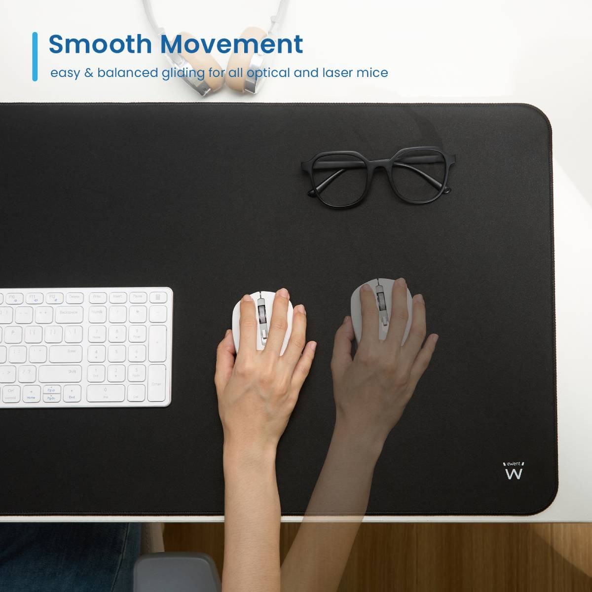 Dłonie używające myszy na czarnej podkładce, klawiatura i okulary na biurku.