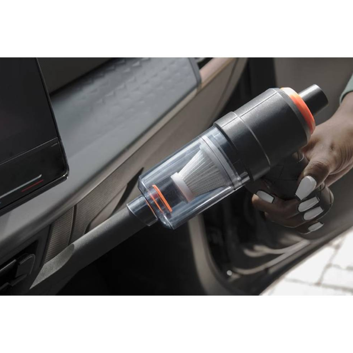 Una persona usa un aspirapolvere portatile in un'auto. È nero e ha un contenitore per la polvere trasparente. L'interno dell'auto è visibile.