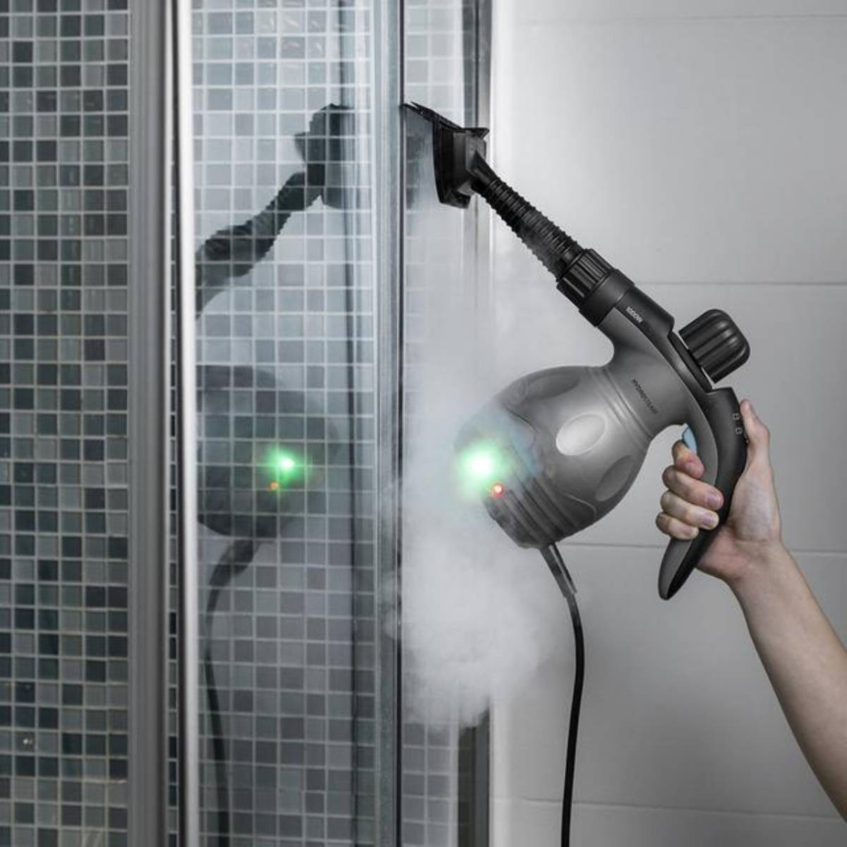 Eine Hand hält einen Dampfreiniger. Dampf wird auf eine Glas-Duschwand freigesetzt. Der Hintergrund ist eine geflieste Wand.