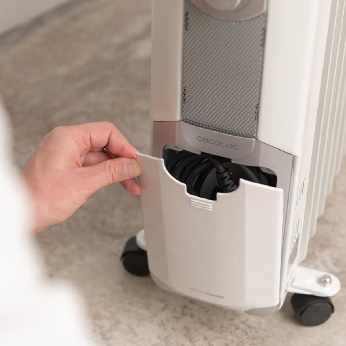 Dłoń osoby otwiera pokrywę, aby odsłonić schowek na przewód zasilający białego grzejnika olejowego.