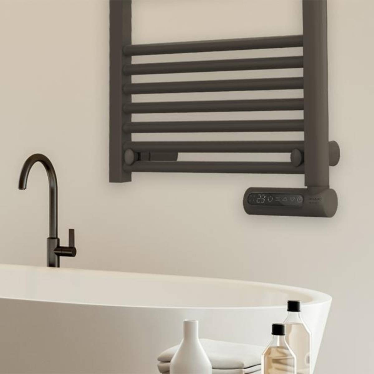 Ciemnoszary podgrzewacz ręczników na białej ścianie. Poniżej biała wanna i czarna bateria. Butelki na pierwszym planie.