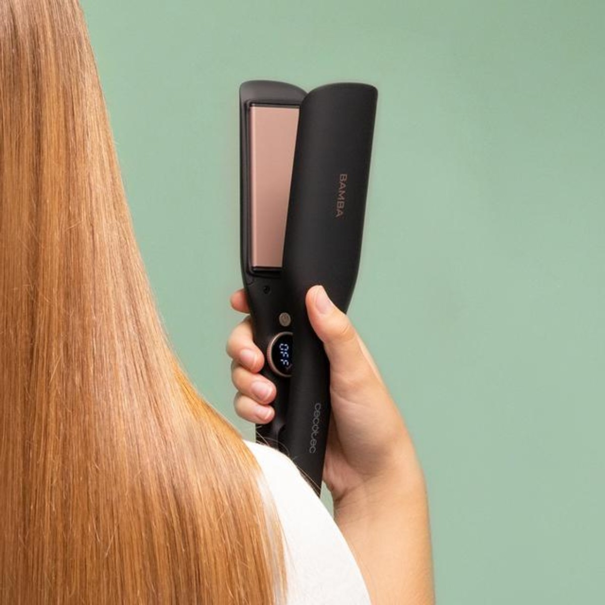 Una persona tiene in mano una piastra nera. Capelli biondi in primo piano e sfondo verde.