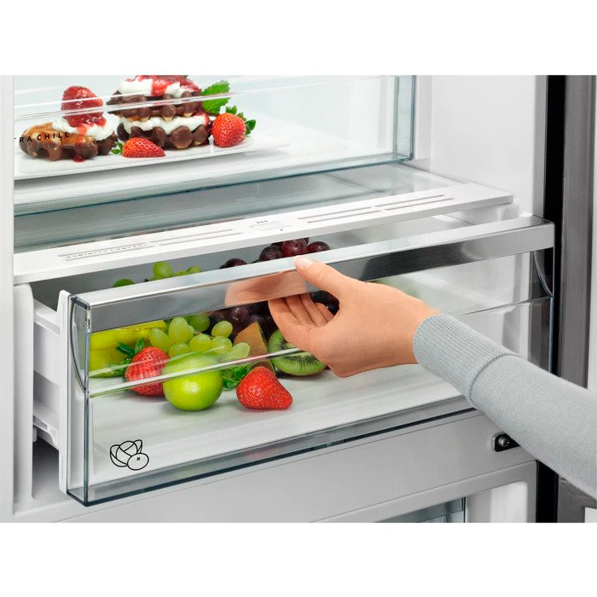 Una mano dentro de un cajón de refrigerador lleno de fruta: fresas, uvas y kiwi.