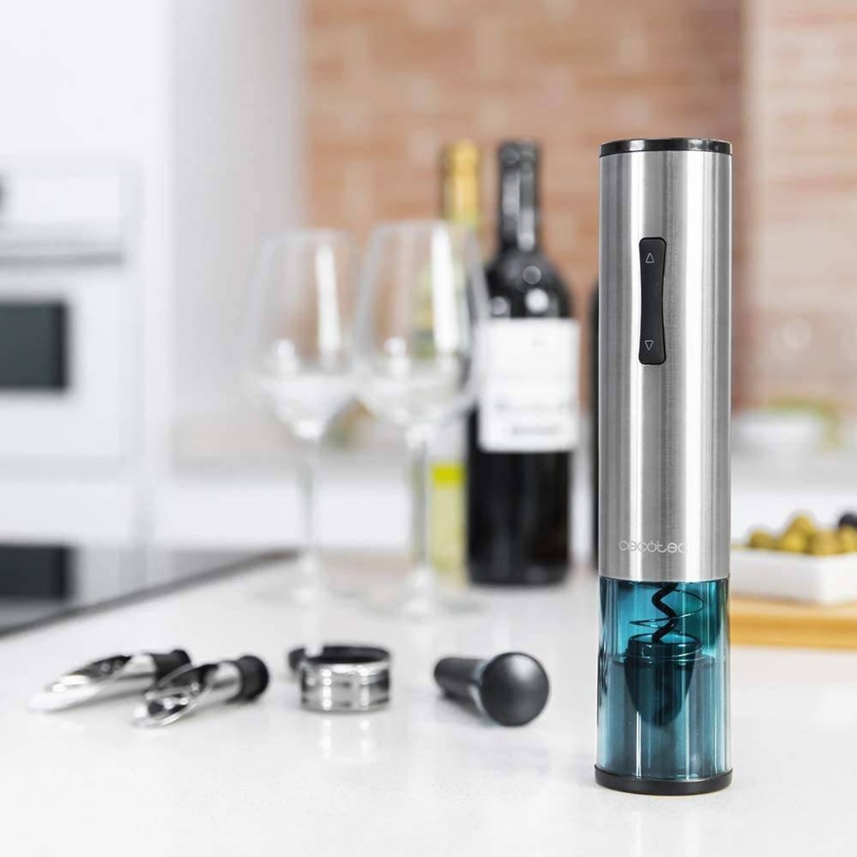 Abridor de vino eléctrico plateado con detalles azules, copas y botellas sobre superficie blanca.