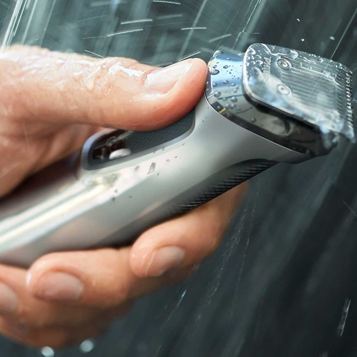 Una mano tiene un rasoio elettrico bagnato, argento e nero sotto l'acqua corrente.