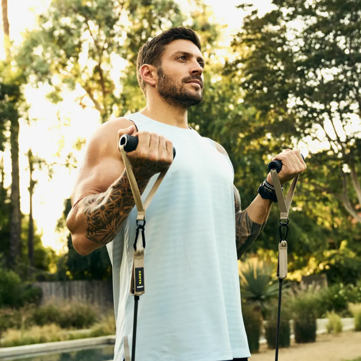 Man exercising with resistance bands outdoors. Light blue tank top and tattoo. Sunny background.